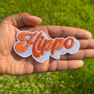 May include: A white iron-on patch with the word "Hippo" embroidered in orange.