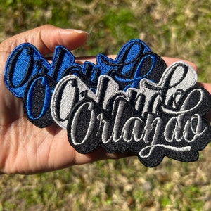 May include: Three embroidered patches featuring the word "Orlando" in a stylised script font. The patches are in blue, white and black.