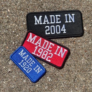 May include: Three rectangular embroidered patches with black borders. The top patch is black with white text reading MADE IN 2004. The middle patch is red with white text reading MADE IN 1982. The bottom patch is blue with white text reading MADE IN 1920.
