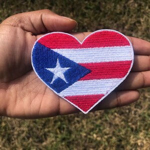 May include: A red, white, and blue heart-shaped patch with the Puerto Rican flag design. The flag features a white star on a blue triangle, with red and white stripes.