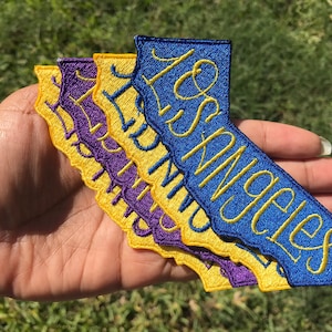 May include: Four embroidered patches in the shape of the state of California. The patches are layered on top of each other. The top patch is blue with yellow lettering that says "Los Angeles". The next patch is yellow with purple lettering that says "Los Angeles". The next patch is purple with yellow lettering that says "Los Angeles". The bottom patch is yellow with purple lettering that says "Los Angeles".