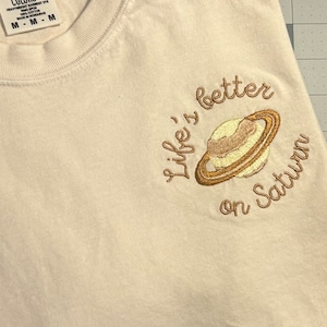 May include: A white t-shirt with a brown embroidered design of a planet with rings and the text "Life's better on Saturn".