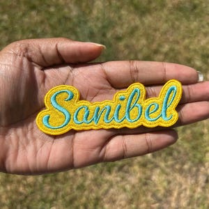 May include: Embroidered patch in a yellow color with the word "Sanibel" in light blue script. The patch has a yellow border and is held in a hand against a blurred green background.