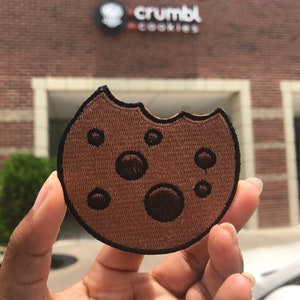 May include: A brown embroidered cookie patch with a bite taken out of it. The patch has five chocolate chips and is in the shape of a cookie. The patch is being held in a hand in front of a brick building with the sign "crumbl cookies" on it.