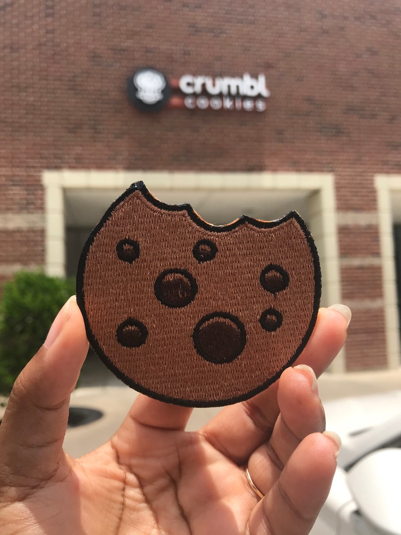 May include: A brown embroidered cookie patch with a bite taken out of it. The patch has five chocolate chips and is in the shape of a cookie. The patch is being held in a hand in front of a brick building with the sign "crumbl cookies" on it.