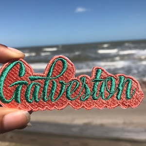 Peut inclure: Un gros plan sur un patch en feutre de couleur corail avec le mot "Galveston" brodé en fil vert. Le patch est tenu dans une main sur un fond flou de plage et d'océan.