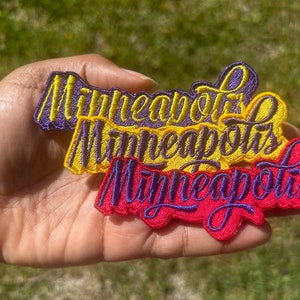 May include: Three embroidered patches with the word "Minneapolis" in a cursive font. The patches are layered on top of each other, with the top patch being purple, the middle patch being yellow, and the bottom patch being red.