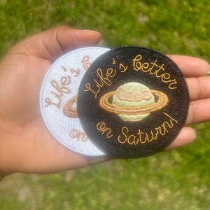 May include: Two embroidered iron-on patches with the text "Life's better on Saturn!" and a depiction of the planet Saturn. One patch is white with gold embroidery, the other is black with gold embroidery.