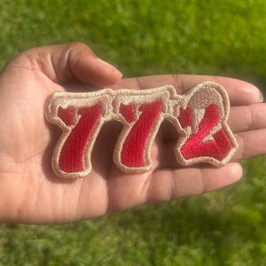 May include: An embroidered patch featuring the numbers 772 in a bold, stylized font. The numerals are filled with a vibrant red thread and outlined with a thick, light beige border that creates a raised, textured appearance against the fabric.