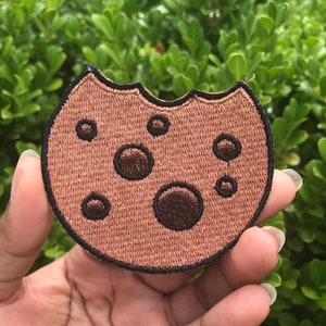May include: A brown embroidered cookie patch with a bite taken out of it. The cookie has chocolate chips and a black outline.