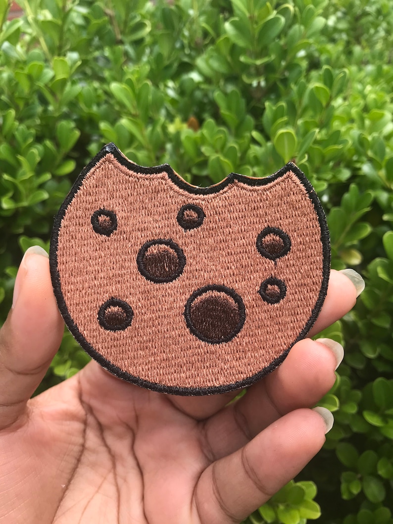 May include: A brown embroidered cookie patch with a bite taken out of it. The cookie has chocolate chips and a black outline.
