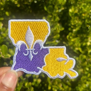 May include: A white, yellow, and purple embroidered patch shaped like the state of Louisiana with a fleur-de-lis in the center.