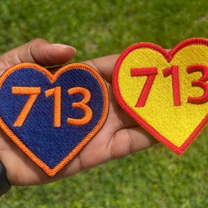 May include: Two heart-shaped patches with the number "713" embroidered on them. One patch is navy blue with orange stitching and numbers, the other is yellow with red stitching and numbers. The patches are likely for clothing or accessories.
