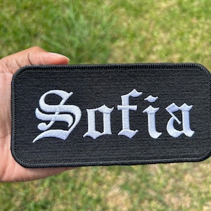 May include: Black rectangular patch embroidered with the name "Sofia" in white gothic lettering. The patch has a black border and is held against a background of green grass.