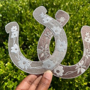 May include: Two embroidered horseshoe patches. The patches are beige and brown, with white floral designs. The patches are intertwined, with a green leafy background.