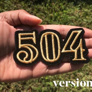 May include: A black embroidered patch with the numbers "504" in gold thread. The patch is held in a hand.