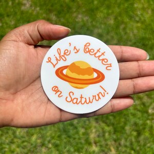 May include: White round sticker with the text "Life's better on Saturn!" in orange cursive lettering. A cartoon image of Saturn with orange rings is in the center of the sticker.
