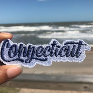 May include: White fabric patch with blue embroidered lettering that reads "Connecticut". The patch is held in a hand against a blurry background of a beach with blue water and white waves.