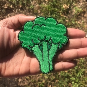 May include: A green embroidered broccoli patch. The patch is in the shape of a broccoli floret with a stem.