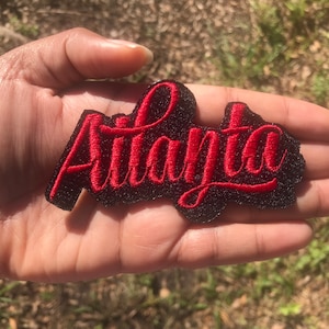 May include: Red embroidered patch with the word "Atlanta" in a cursive font on a black background.