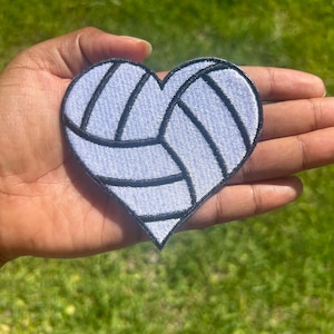 Peut inclure: Un patch en forme de cœur blanc avec un motif de volley-ball noir. Le patch est brodé avec un fond blanc et des points noirs.