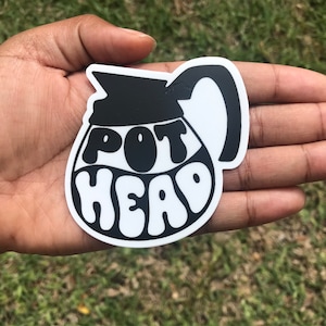 May include: A black and white sticker in the shape of a coffee pot. The sticker has the words "POT HEAD" in a retro font. The sticker is held in a hand against a background of green grass.