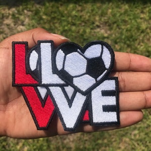 May include: A set of three iron-on patches featuring the word "LOVE" with a soccer ball in the shape of a heart. The patches are red, white, and black.
