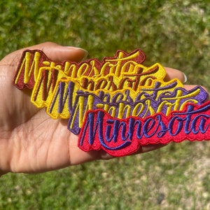 May include: Four embroidered patches with the word "Minnesota" in different colors. The patches are stacked on top of each other, with the red patch on the bottom and the yellow patch on the top. The patches are all in a cursive font.