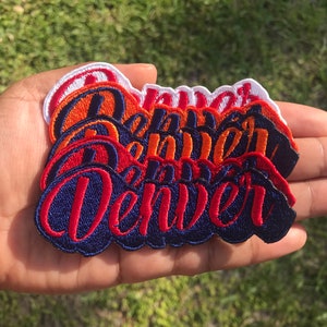 May include: A stack of embroidered iron-on patches with the word "Denver" in a stylized font. The patches are in various shades of red, orange, and blue.