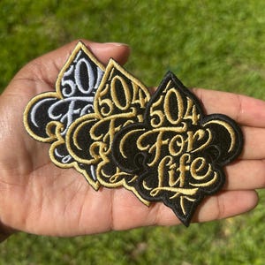 May include: Three embroidered patches shaped like fleur-de-lis symbols. Two patches feature gold thread embroidery on a black background, while one patch features silver thread embroidery on a black background. Each patch displays the text 504 For Life.