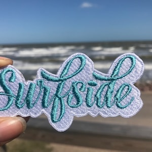 May include: A white embroidered patch with the word "Surfside" in teal blue lettering. The patch is held in a hand against a blurry background of a beach and ocean.