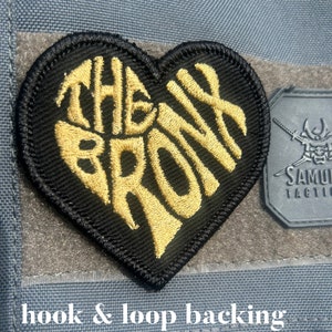 May include: A heart-shaped embroidered patch with a black background and gold stitching that reads THE BRONX. The patch features a hook and loop backing and measures 3.2 inches by 3.2 inches. It is attached to a grey fabric surface.