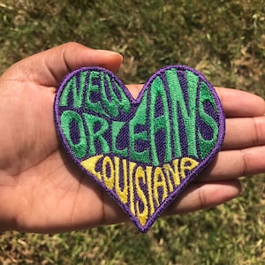 May include: A purple heart-shaped embroidered patch with the words "New Orleans Louisiana" in green and yellow lettering.