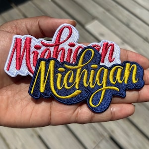 Peut inclure: Deux écussons brodés avec le mot "Michigan" en écriture cursive. L'un a des lettres rouges sur fond blanc, l'autre a des lettres jaunes sur fond bleu marine.