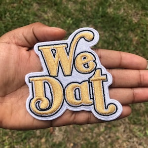 May include: Embroidered patch with the words "We Dat" in gold lettering with a black outline on a white background. The patch is held in a hand against a backdrop of green grass.