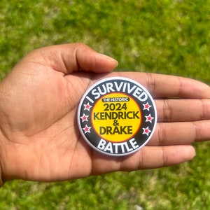 May include: A black and white button with the text "I Survived The Historic 2024 Kendrick & Drake Battle" in white lettering on a yellow background. The button is surrounded by six red stars.