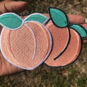 May include: Two embroidered peach patches. The peaches are a light orange color with a white border and green leaves. One patch has a black outline, and the other has a white outline. The patches are likely for clothing or accessories.