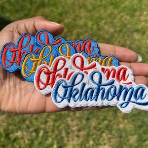 May include: A collection of embroidered patches featuring the word Oklahoma in a cursive script. The patches are available in various color combinations, including red text on blue, yellow text on blue, red text on white, and blue text on white.