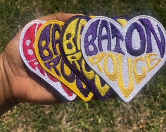 May include: Four heart-shaped patches with the text "BATON ROUGE" in a purple and yellow color scheme. The patches are arranged in a stack, with the largest patch on top and the smallest patch on the bottom.