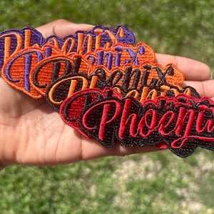 May include: A stack of embroidered iron-on patches with the word "Phoenix" in different colors, including purple, orange, black, and red.