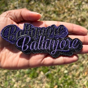 Peut inclure: Deux patchs brodés violets et noirs avec le mot "Baltimore" en écriture cursive.