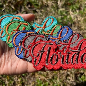 May include: A set of six embroidered iron-on patches featuring the word "Florida" in a cursive script. The patches are in various colors, including red, green, blue, and turquoise, with black outlines.