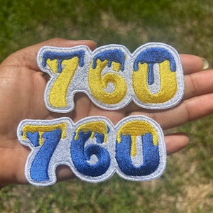 Peut inclure: Deux patchs thermocollants avec le numéro 760 dans une police d'écriture qui coule. Le patch du haut est jaune avec des gouttes bleues, et le patch du bas est bleu avec des gouttes jaunes.