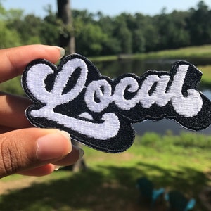 May include: White embroidered "Local" patch on a black background. The patch is being held by a hand.