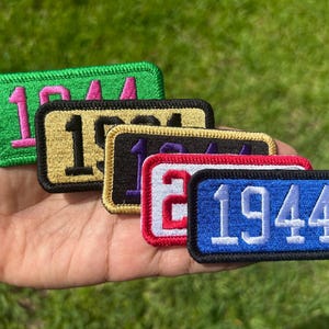 May include: A collection of five rectangular embroidered patches, each featuring the number 1944. The patches come in various color combinations, including green with pink, gold with black, black with purple, white with red, and blue with white.