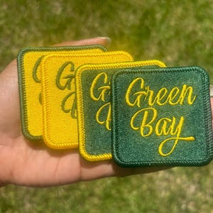 May include: Four iron-on patches, two yellow and two green. The yellow patches say "Green Bay" in a cursive font. The green patches say "Green Bay" in a cursive font.