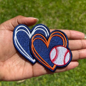 May include: Embroidered patches in the shape of hearts and a baseball. The patches are navy blue with white and orange accents. The baseball has red stitching. The patches are held in a hand.