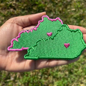 May include: Two green felt patches in the shape of the state of Kentucky, with pink outlines and pink hearts in the center. The patches are held in a person's hand.