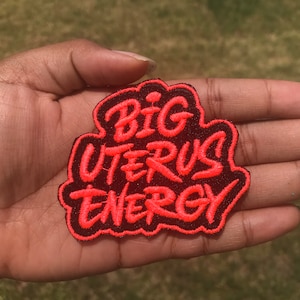May include: A black and red embroidered patch with the text "BIG UTERUS ENERGY" in a bold, stylized font.