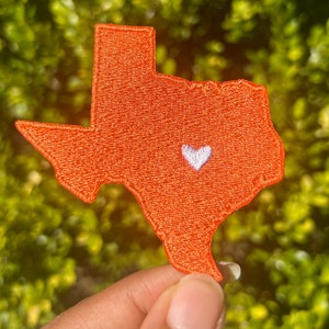 May include: An orange embroidered patch in the shape of the state of Texas, featuring a white heart in the center. The patch is held against a blurred green background, highlighting the detailed stitching.